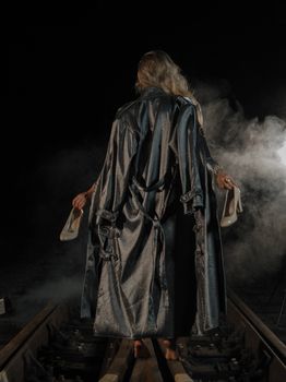 A woman in a stylish robe walks barefoot on tracks at night, holding shoes amidst smoke.