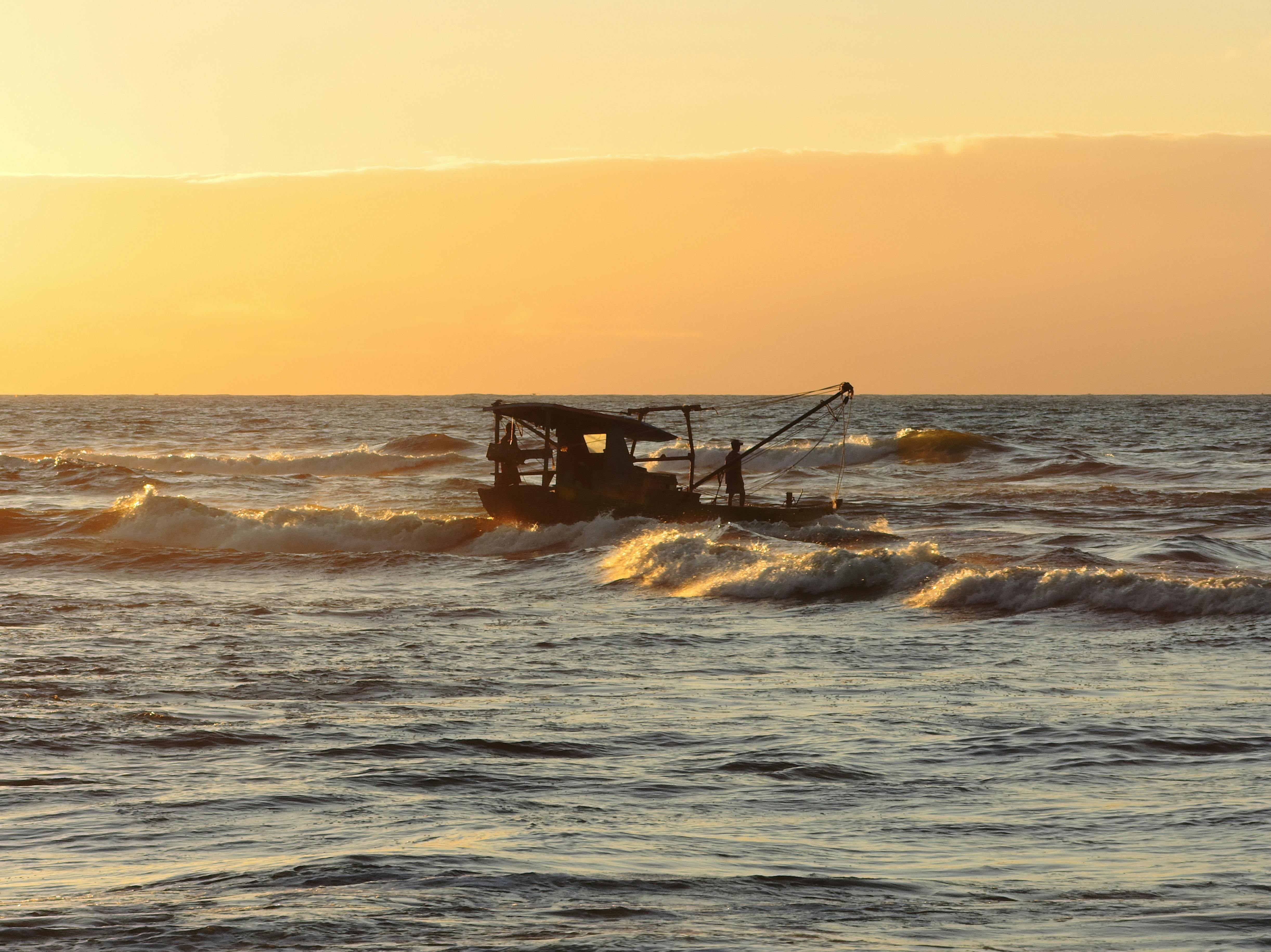 Free A fishing boat silhouettes against a vibrant orange sunset on the ocean, capturing the tranquil beauty of the sea. Stock Photo