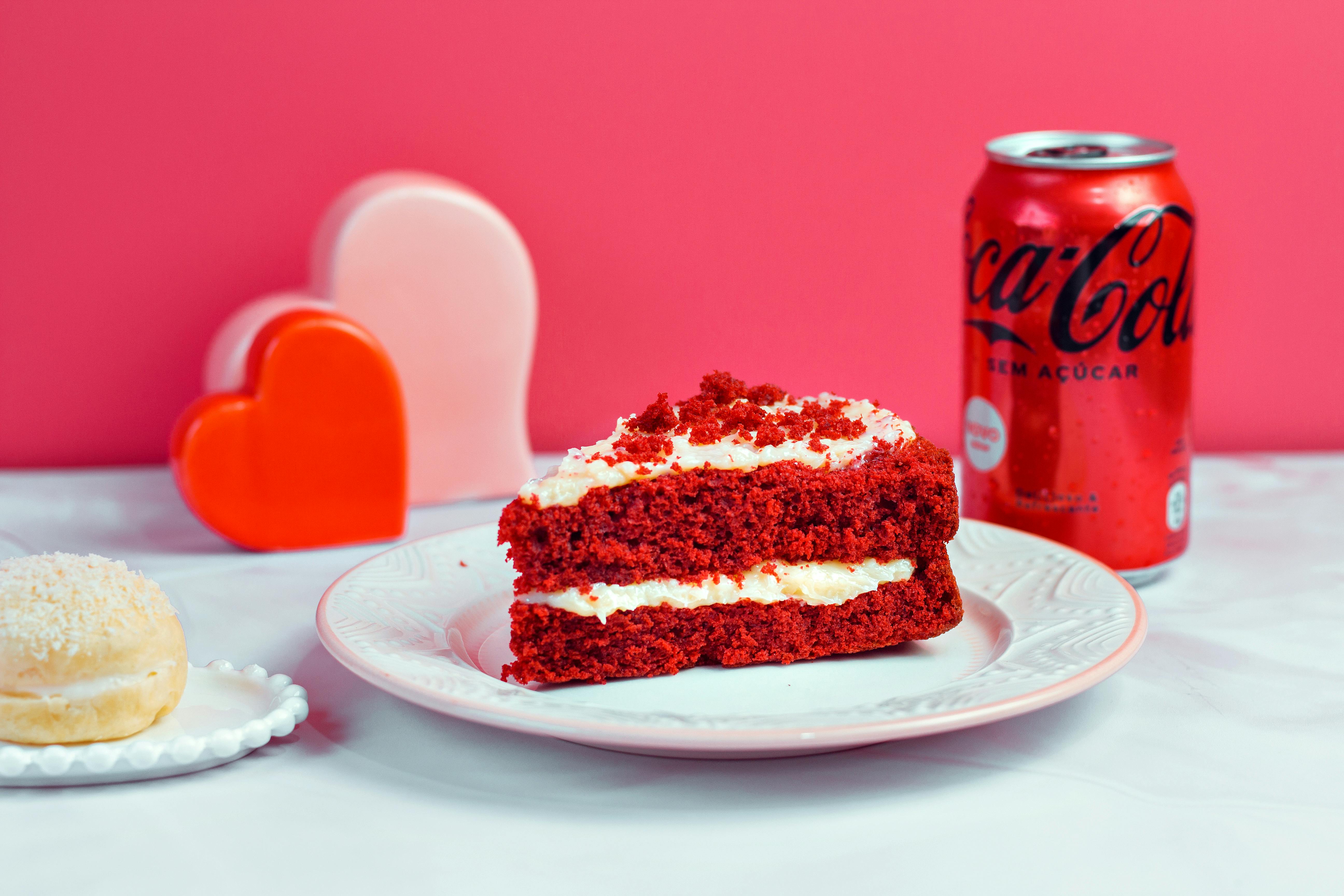 Slice of red velvet cake on ceramic plate with coke on pink background.