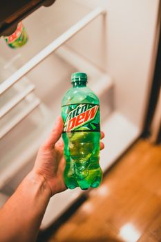 Close-up of a hand holding a Mountain Dew bottle in front of an open refrigerator.