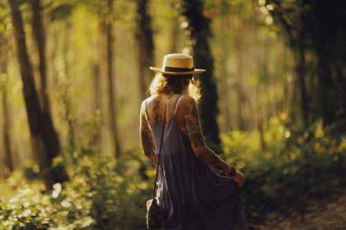 Free A woman in a hat explores a sunlit forest, showcasing tattoos and a flowing dress. Stock Photo