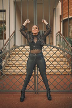 Fashionable woman in black leather pants leans against a metal gate outdoors.