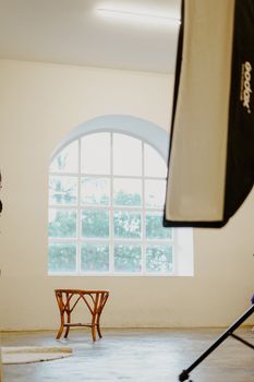 Bright studio with large window, featuring a rattan stool and professional lighting equipment. - Fotografer Sidoarjo
