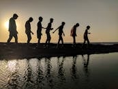 一群孩子沿着海岸线行走，日落映衬着他们的轮廓，倒影在水中