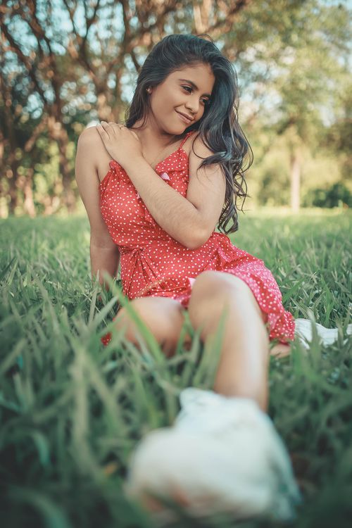 Free Portrait of a young woman in a polka dot dress sitting on grass, enjoying a sunny day. Stock Photo