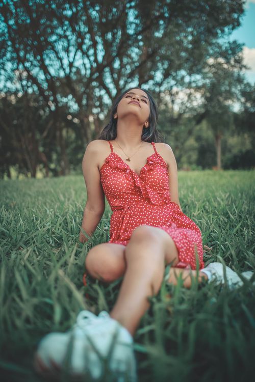 Free Young woman sitting outdoors on grass in a red polka dot dress enjoying a peaceful moment. Stock Photo
