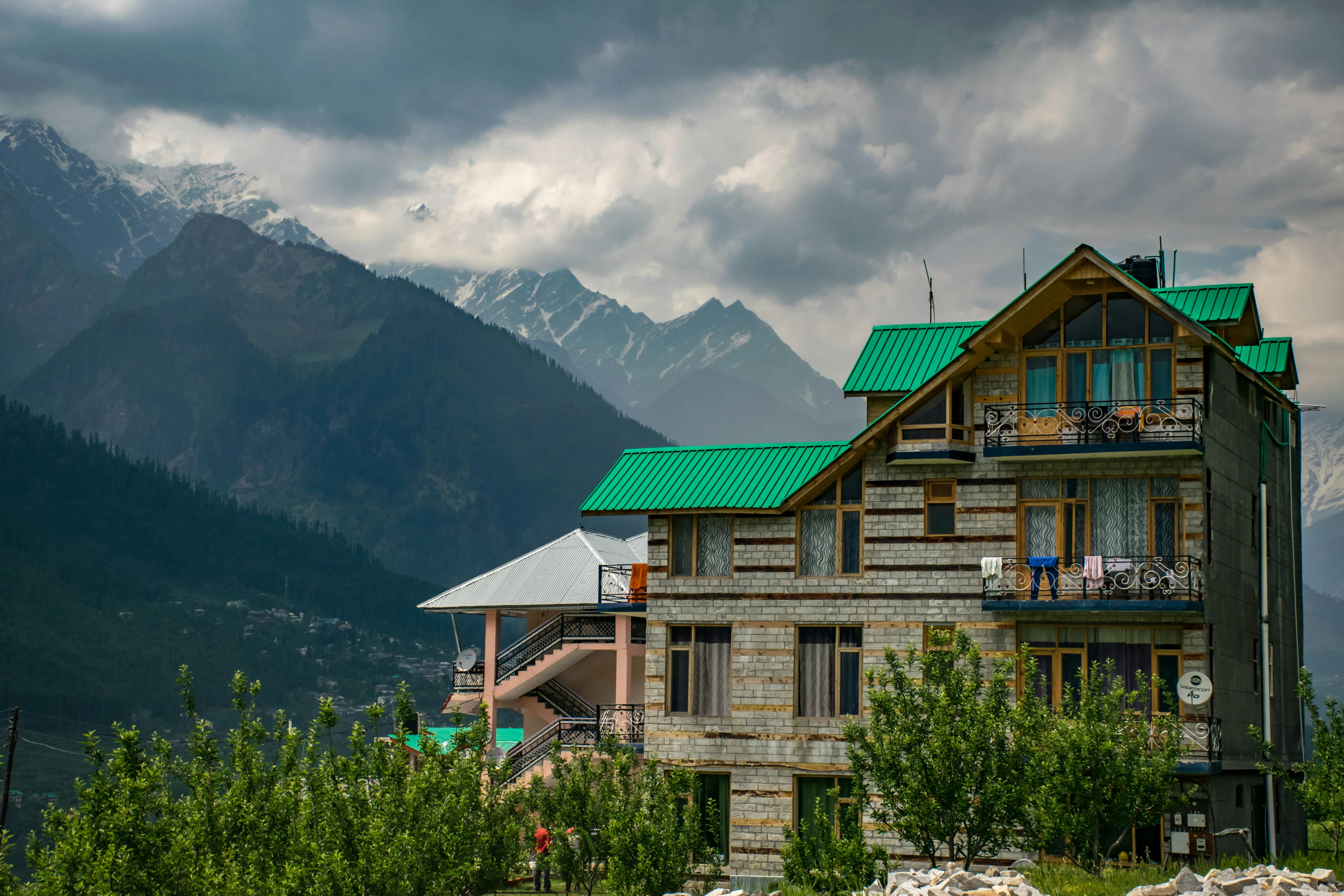 Modern mountain house with green roof, set against snow-capped peaks under cloudy skies.