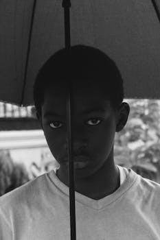 Black and white close-up portrait of a young boy standing under an umbrella, expressing innocence and introspection.