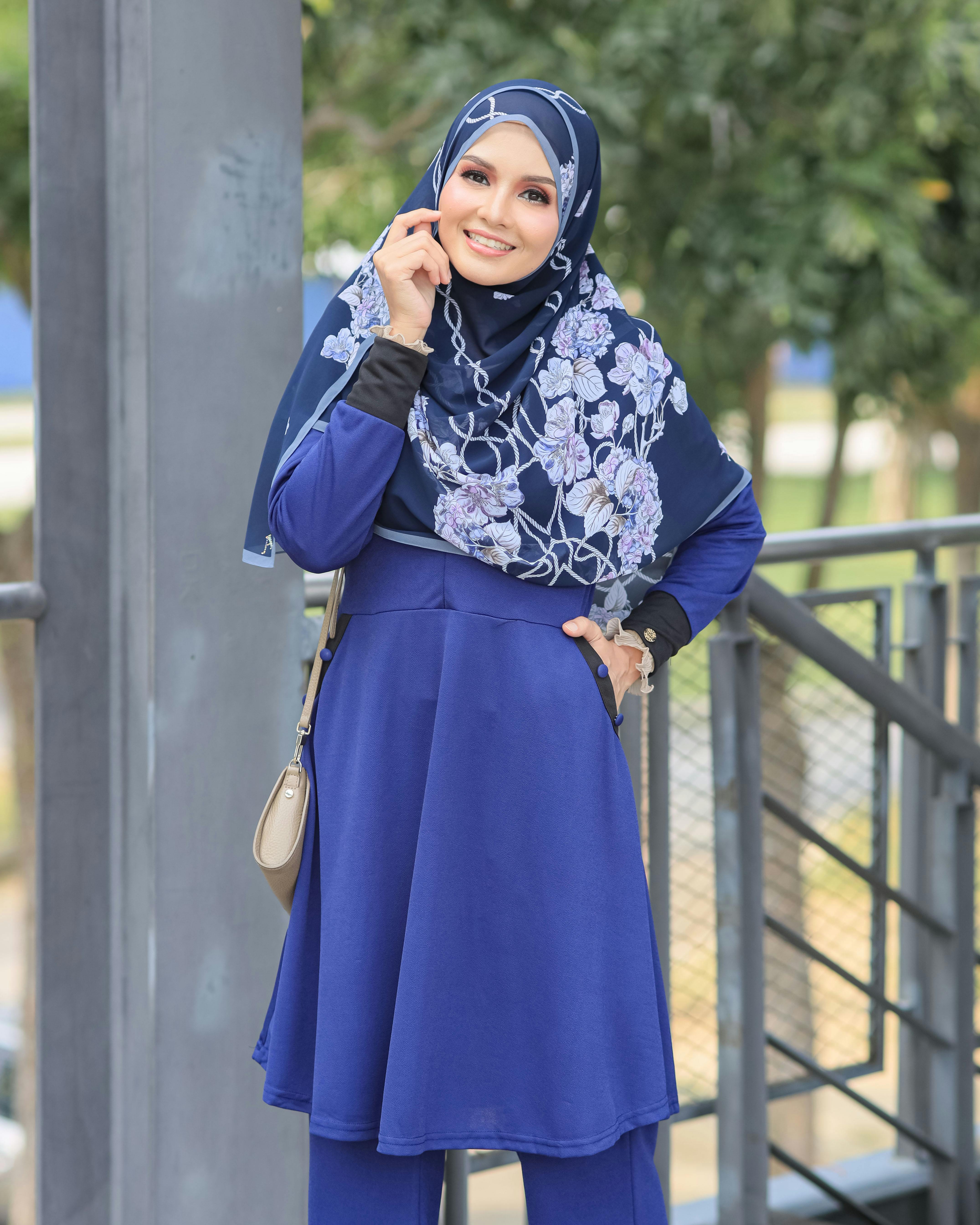 A cheerful woman in a hijab poses outdoors in a stylish blue dress.