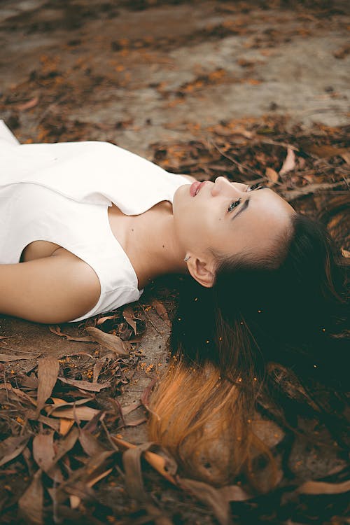 Free A Woman in White Tank Top Lying on the Ground with Brown Dried Leaves Stock Photo