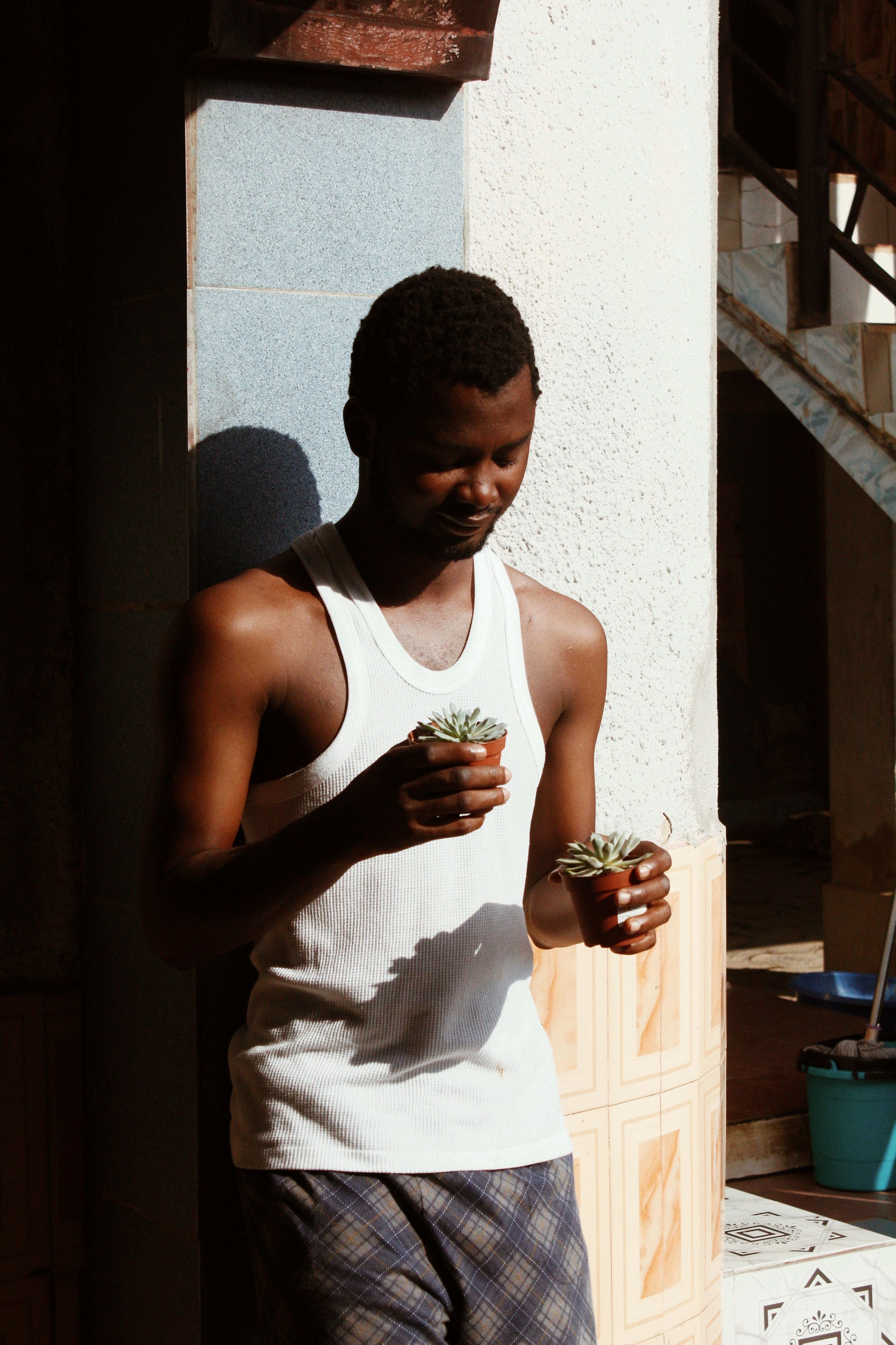 A relaxed man standing indoors, holding potted succulents, enjoying a sunlit moment.