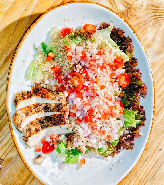 A wide bowl layered with quinoa, sliced grilled chicken, cucumber, cherry tomatoes, red onion, kalamata olives, and crumbled feta, drizzled with yogurt sauce.
