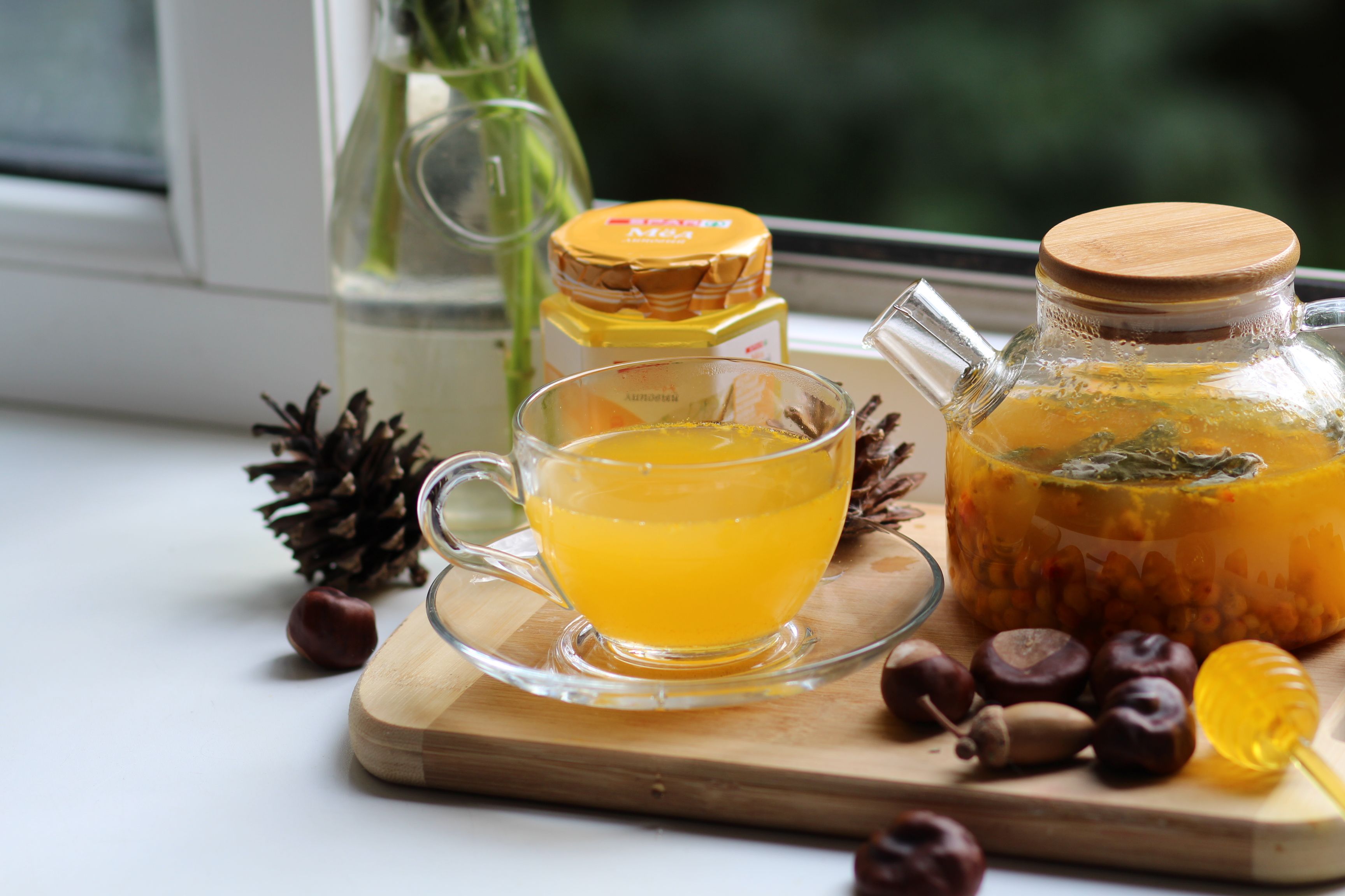 Warm herbal tea in a glass cup with honey, chestnuts, and pinecones on a wooden tray, cozy indoor setting.