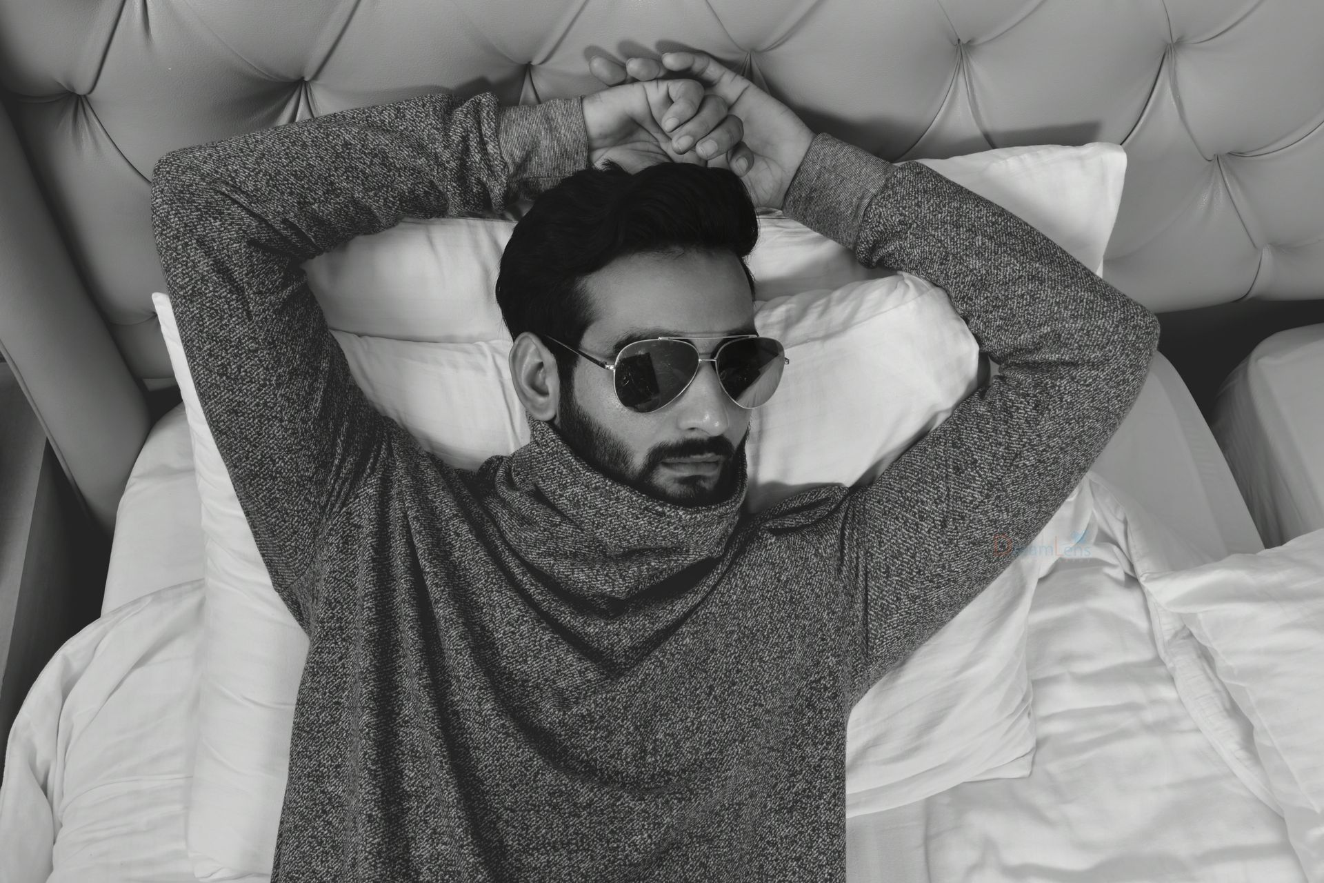 Stylish male model lying on bed wearing sunglasses in a monochrome room.