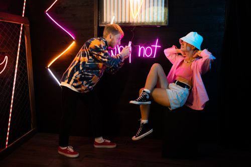 Free Stylish photoshoot with neon lights, featuring a man taking photos of a fashionable woman posing. Stock Photo