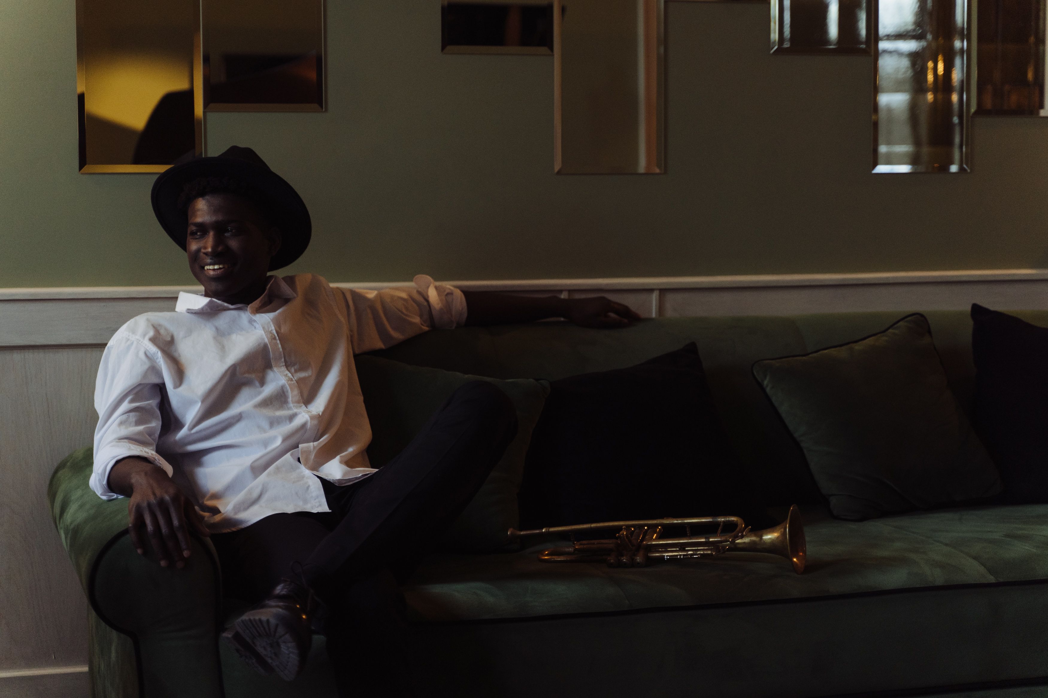 A man in a hat sits on a couch, smiling next to a trumpet indoors.