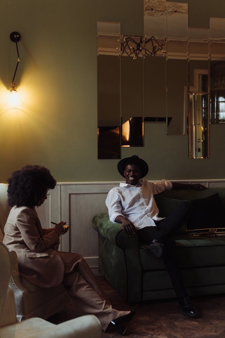 Woman Sitting On White Chair Doing An Interview With Man In White Dress Shirt And Fedora Hat