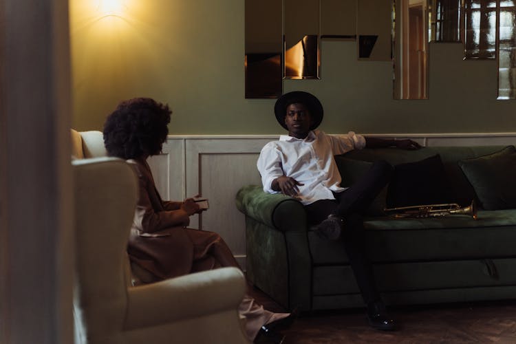 Woman Sitting On White Chair Doing An Interview With Man In White Dress Shirt And Fedora Ha