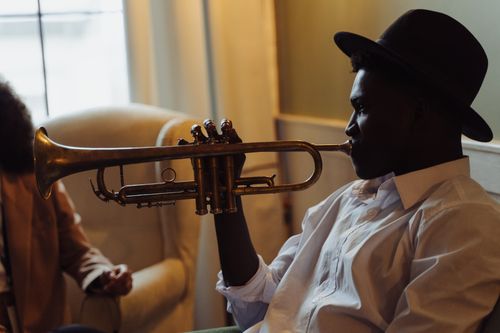 Free A musician in a fedora hat plays the trumpet while seated indoors, creating a classic feel. Stock Photo