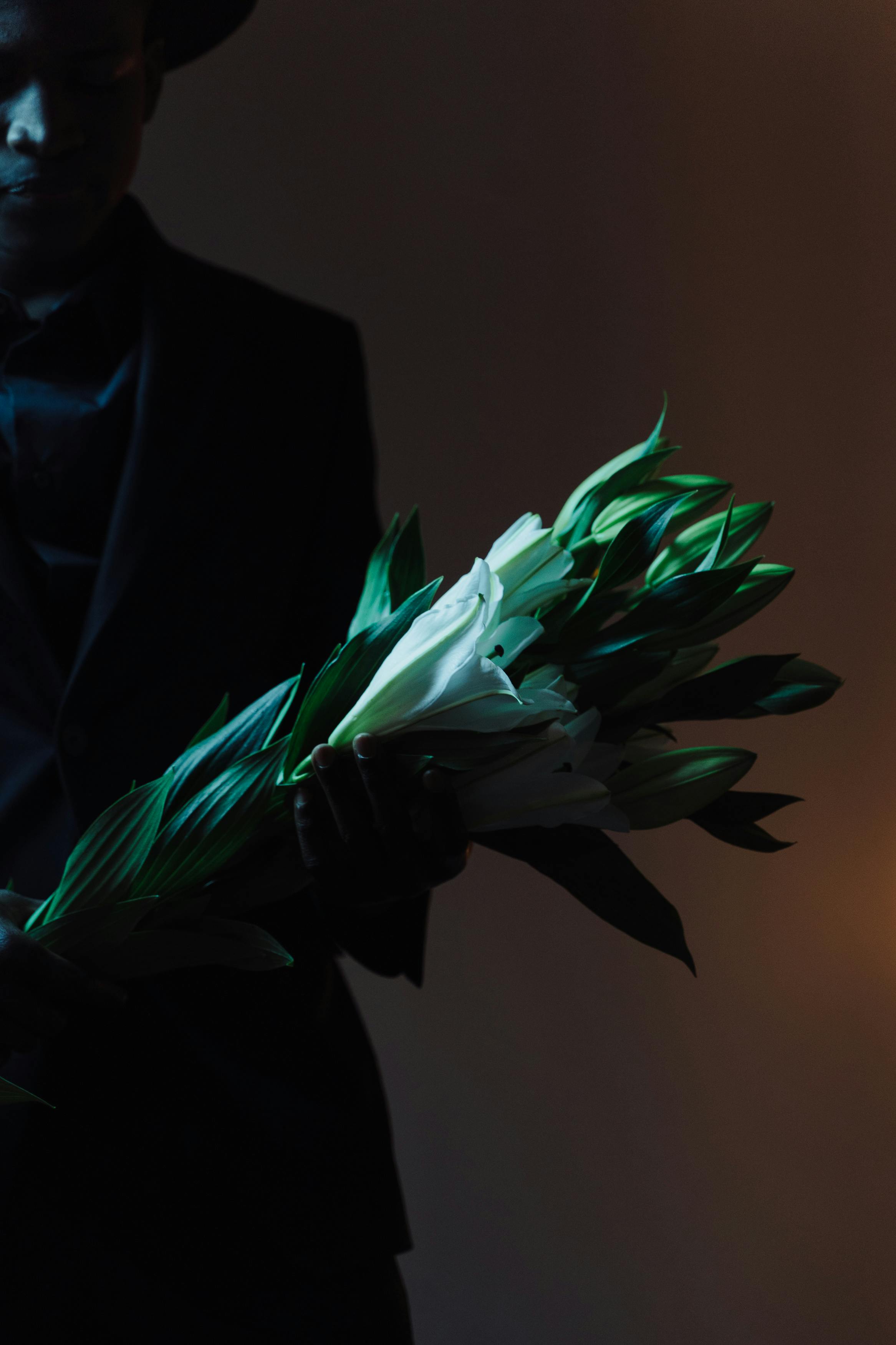 Stylish man in dark suit holding a bouquet of white lilies against a dark backdrop.
