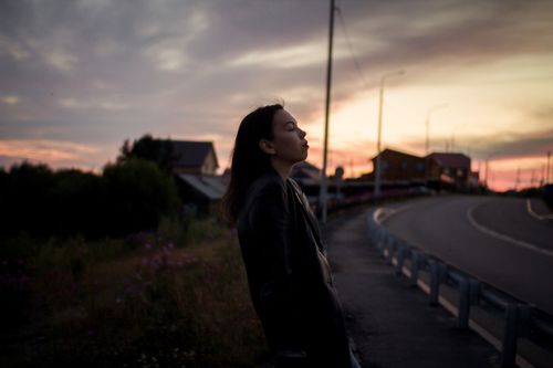 Free Silhouetted woman in a black coat leaning on railings by a rural road during sunset. Stock Photo