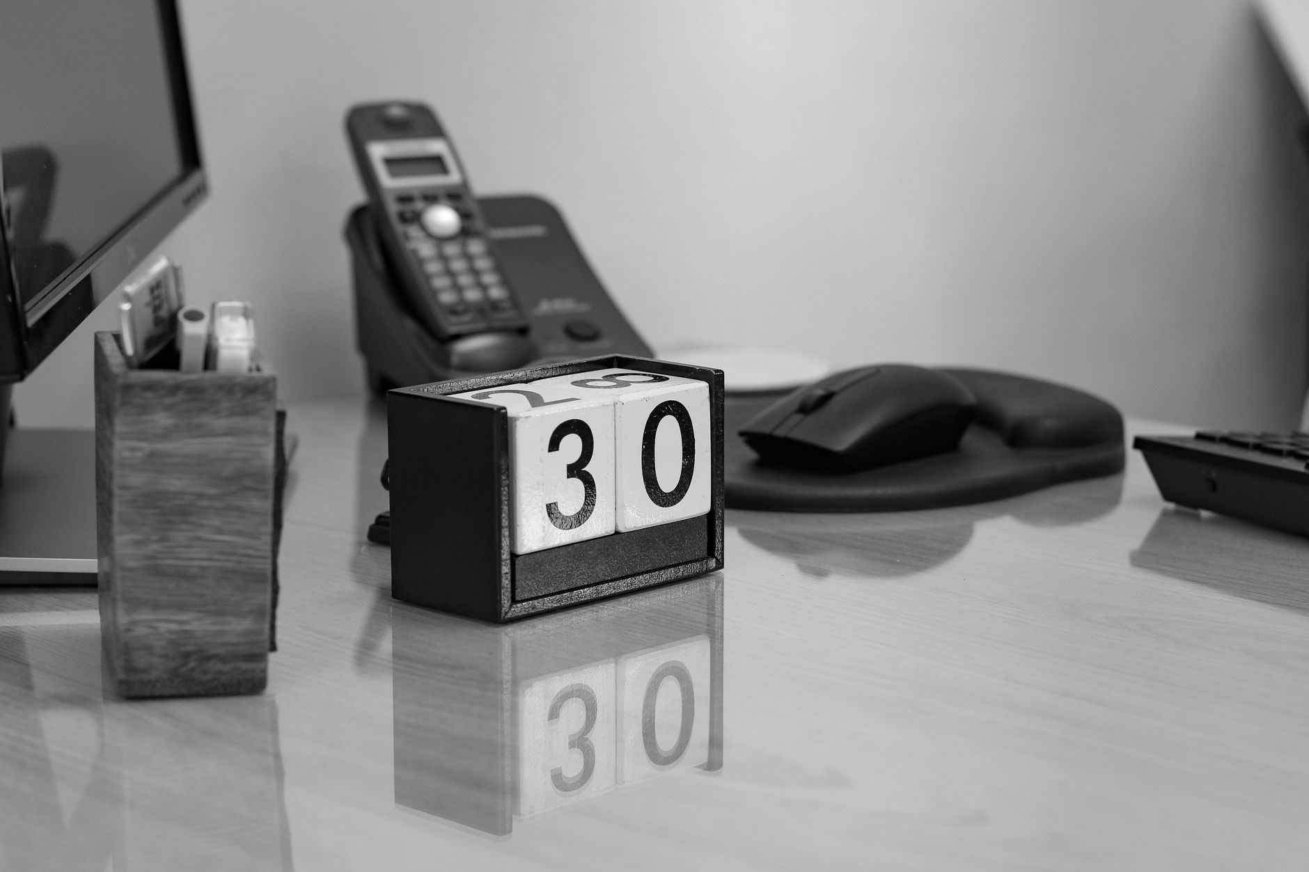 Monochrome image of an office desk with a perpetual calendar, phone, and monitor.