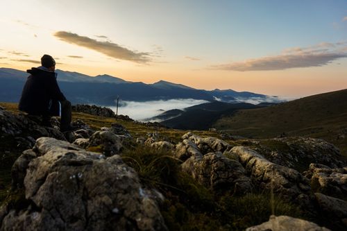 Free A lone hiker enjoys a tranquil sunset on a rocky mountain peak with clouds below. Stock Photo