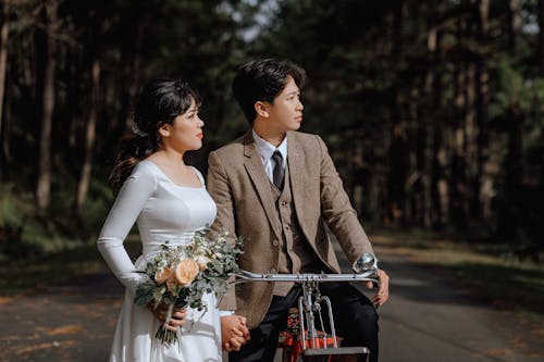 Man in Brown Suit Sitting on a Bike Beside Woman in White Wedding Dress Holding Bouquet of Flowers