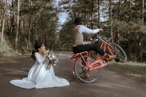 Man in Brown Suit Sitting on a Bike Beside Woman in White Wedding Dress Holding Bouquet of Flowers