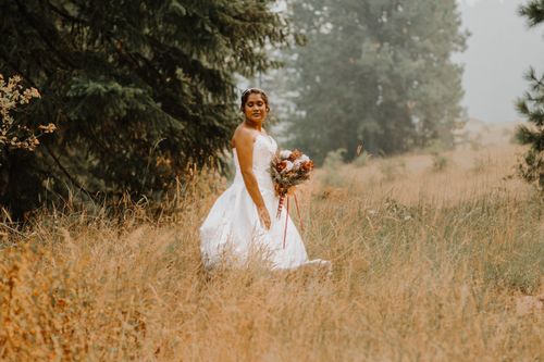 Free An elegant bride in a wedding dress holds a bouquet in a scenic hay field, creating a serene outdoor wedding portrait. Stock Photo