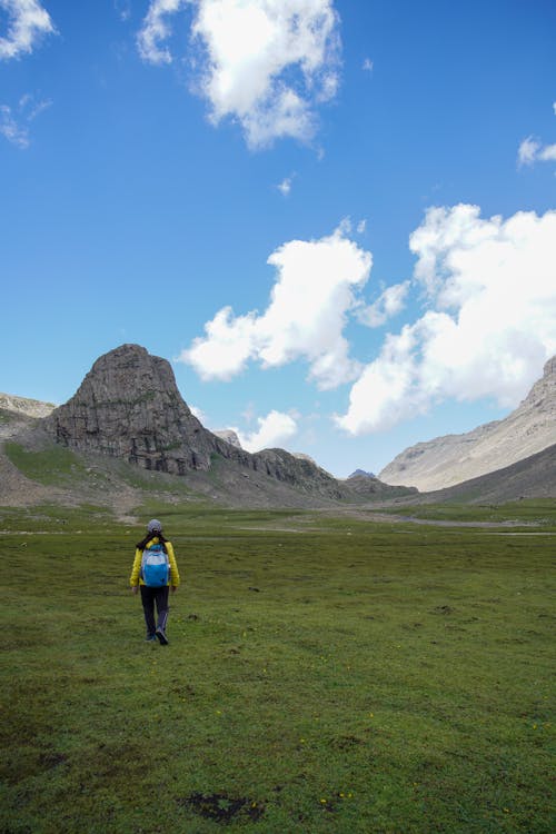 Person in Yellow Jacket and Black Pants Walking on Green Grass Field Near Mountain Under Blue Sky