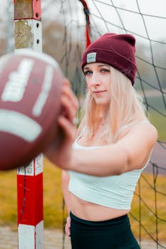 A woman in a beanie holds a football near a soccer goal outdoors.