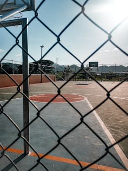 A deserted outdoor basketball court viewed through a chain link fence under clear sky.