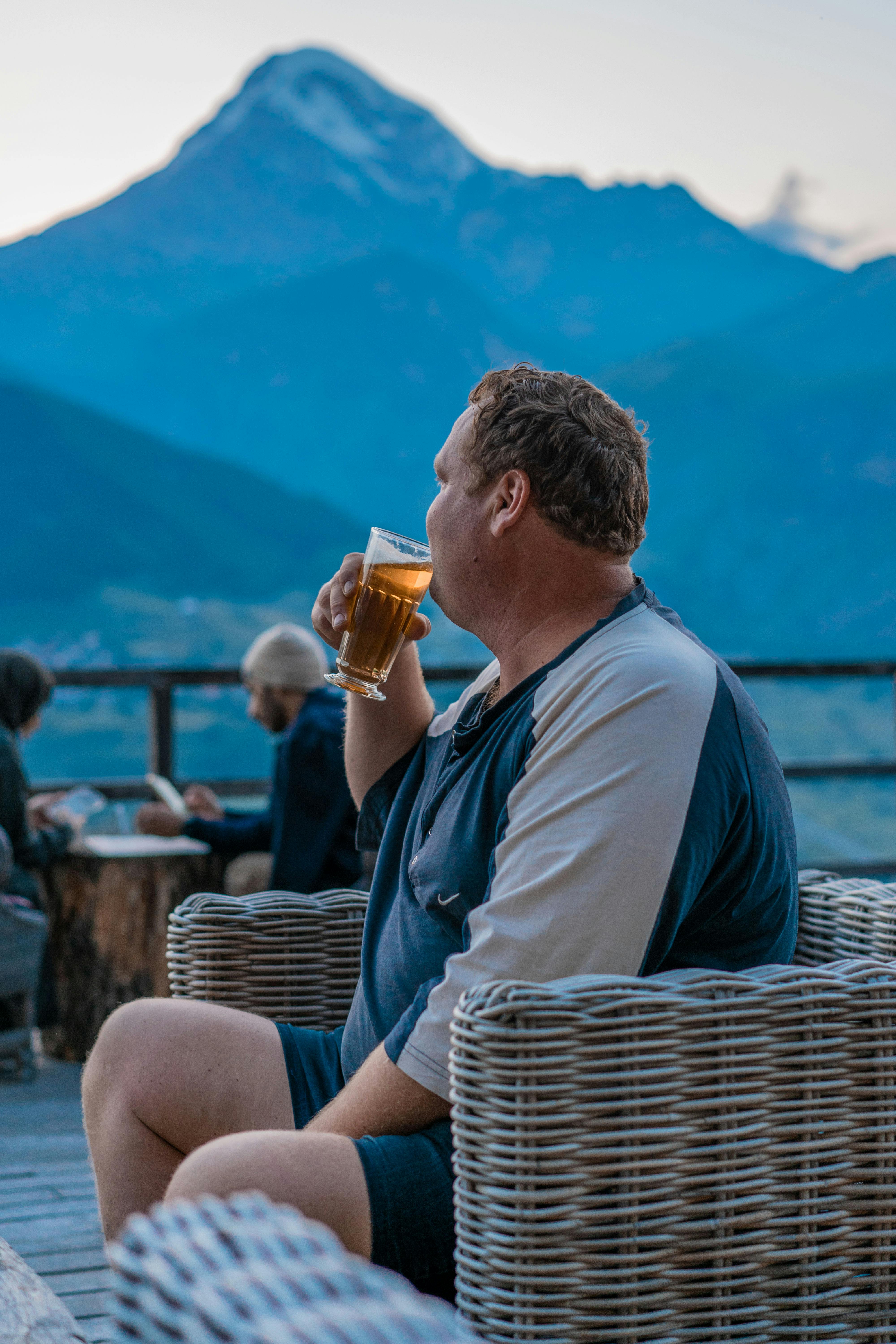 A man relaxes in a wicker chair, sipping beer with a scenic mountain backdrop.