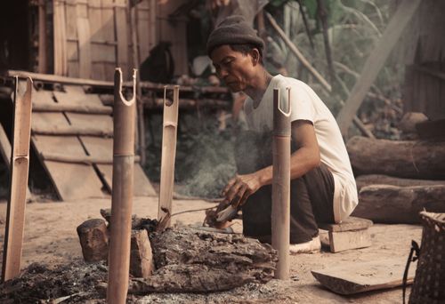 Free An elderly man sitting outdoors, preparing bamboo over an open bonfire in a rural setting. Stock Photo