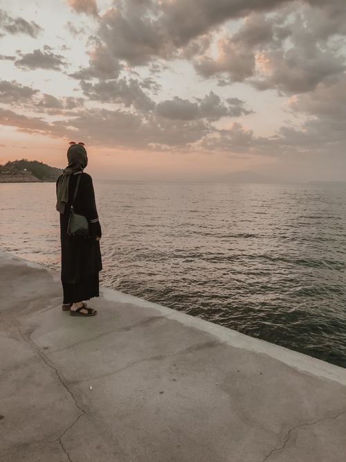 Free A woman in a black robe and headscarf stands on a pier, gazing at the ocean during a serene sunset. Stock Photo