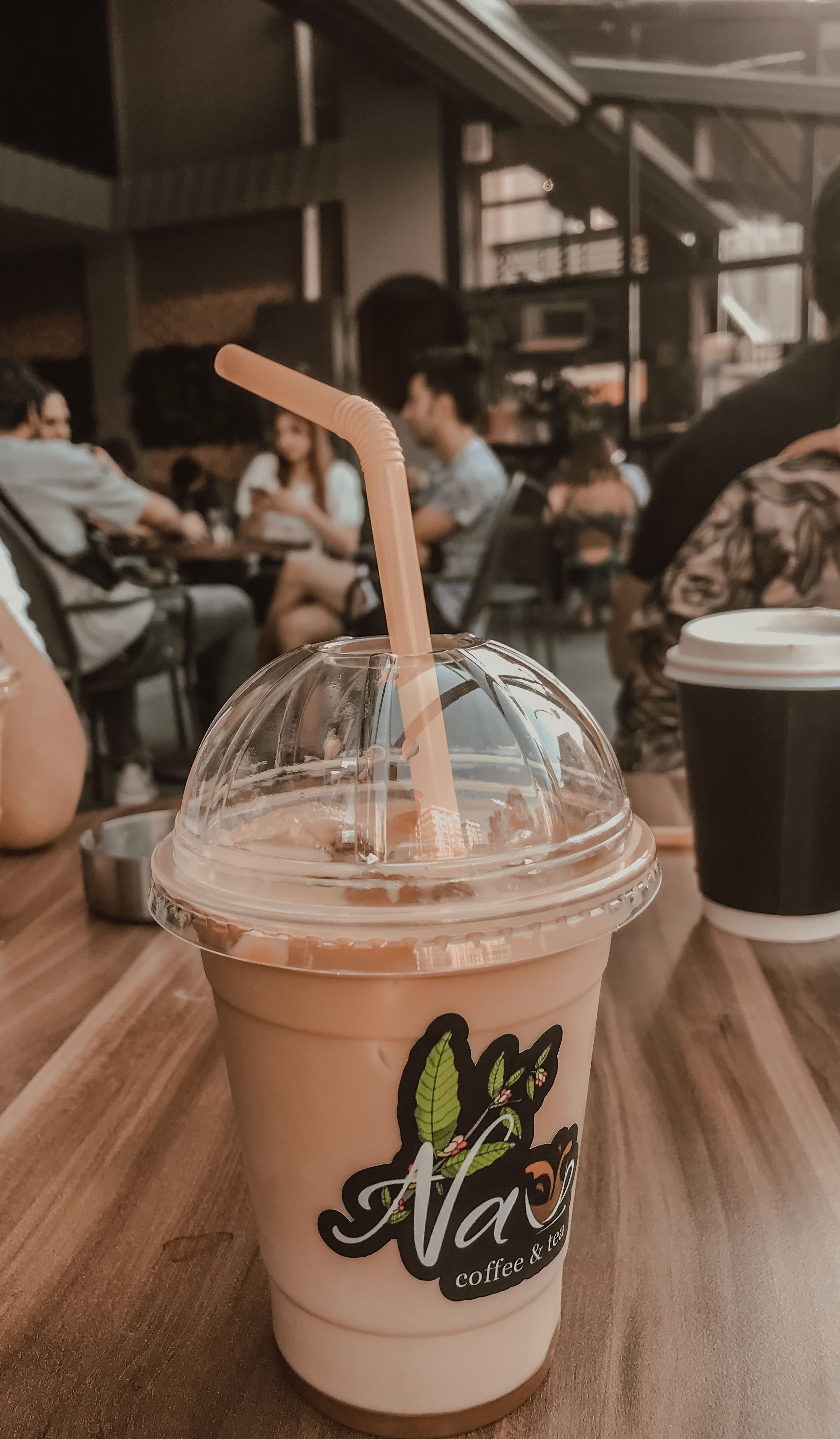 A refreshing iced coffee with straw on a wooden table in a busy cafe.