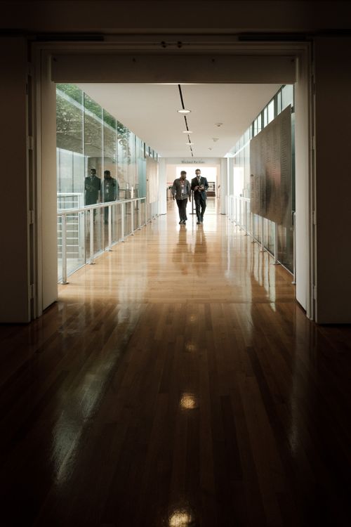Free Three business colleagues walking together in a bright, modern office hallway with large windows and wooden flooring. Stock Photo