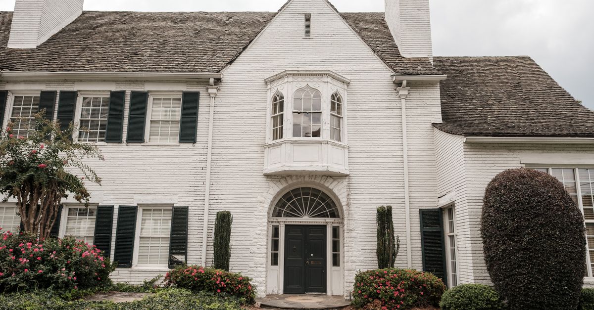 Elegant white brick house with green shutters, lush garden, and a curved walkway accentuating its classic facade.
