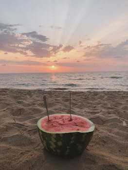 A slice of watermelon placed on sandy beach with stunning sunset view over the sea.