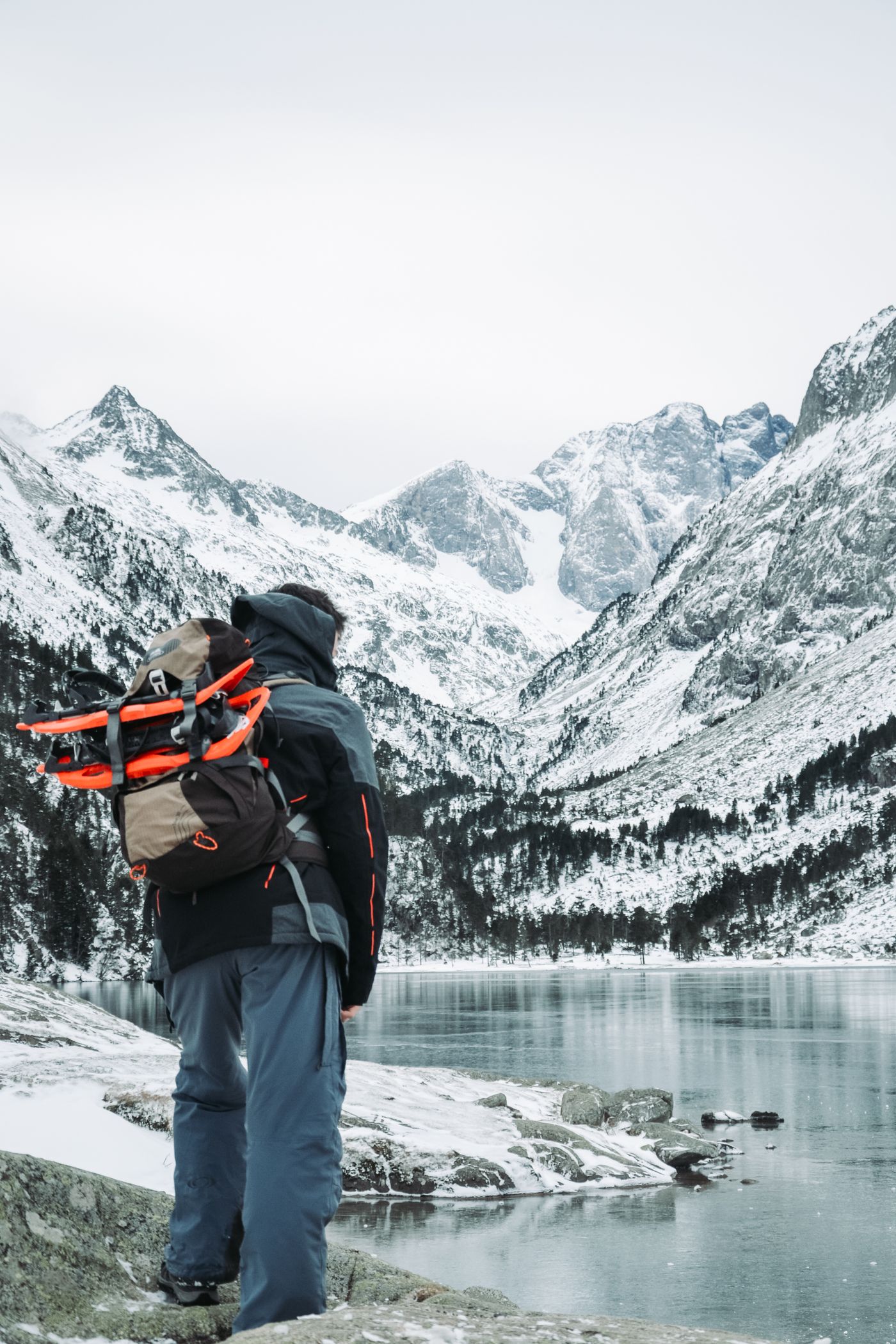 A hiker carrying a backpack standing by a frozen lake in snowy mountains