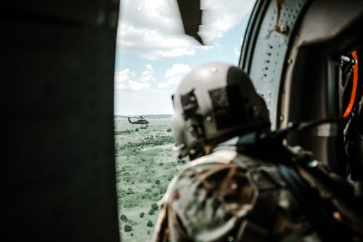 Helmeted soldier in helicopter observing another helicopter over a vast landscape.