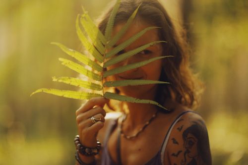 Free Close-up of a woman playfully hiding behind a fern leaf in a sunlit forest setting. Stock Photo