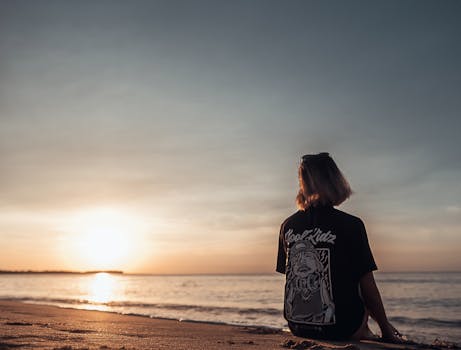 Woman sitting on a beach enjoying a serene sunrise over the ocean, embracing nature and tranquility.