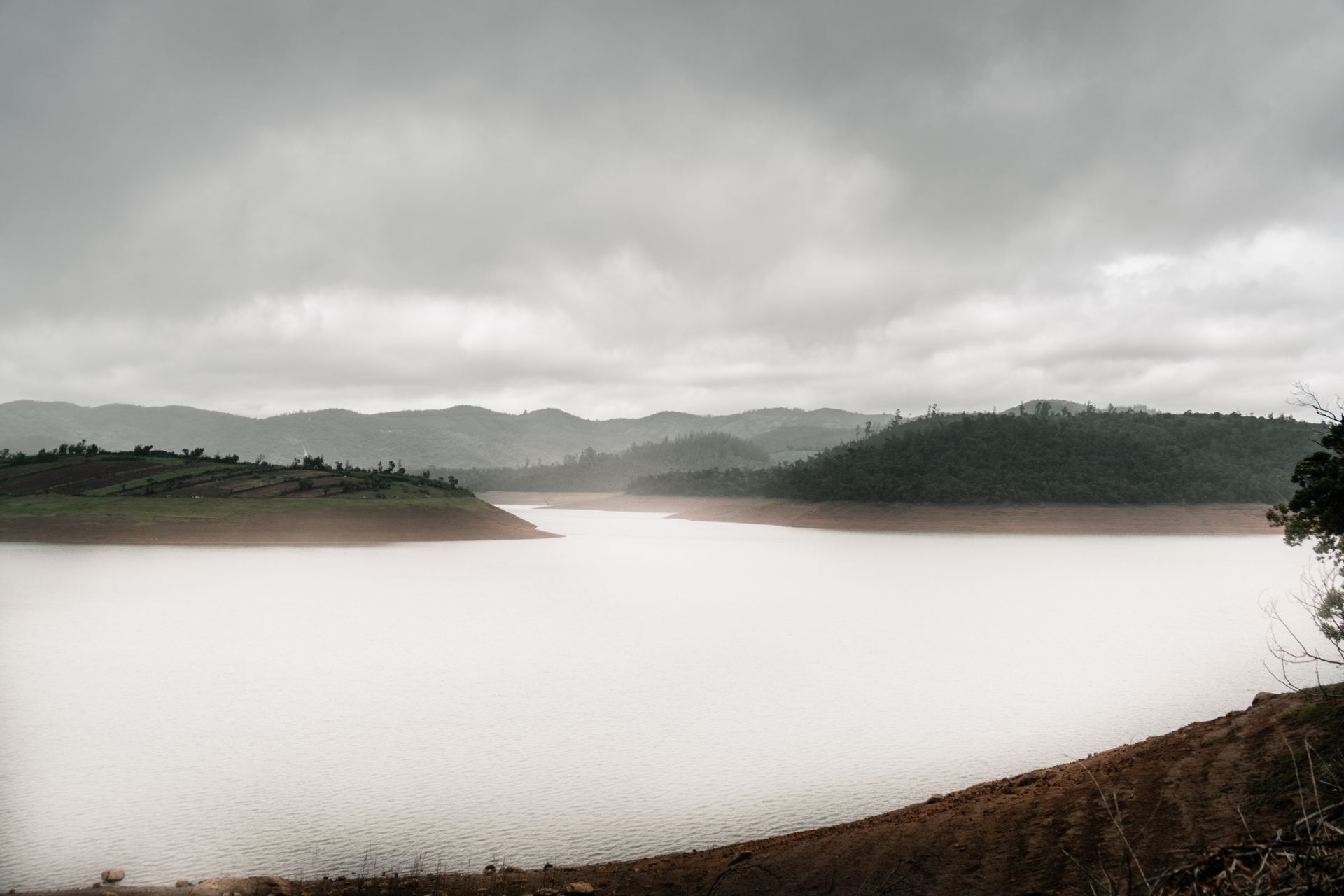 Serene view of a lake surrounded by hills and fog under an overcast sky in Tamil Nadu, India.