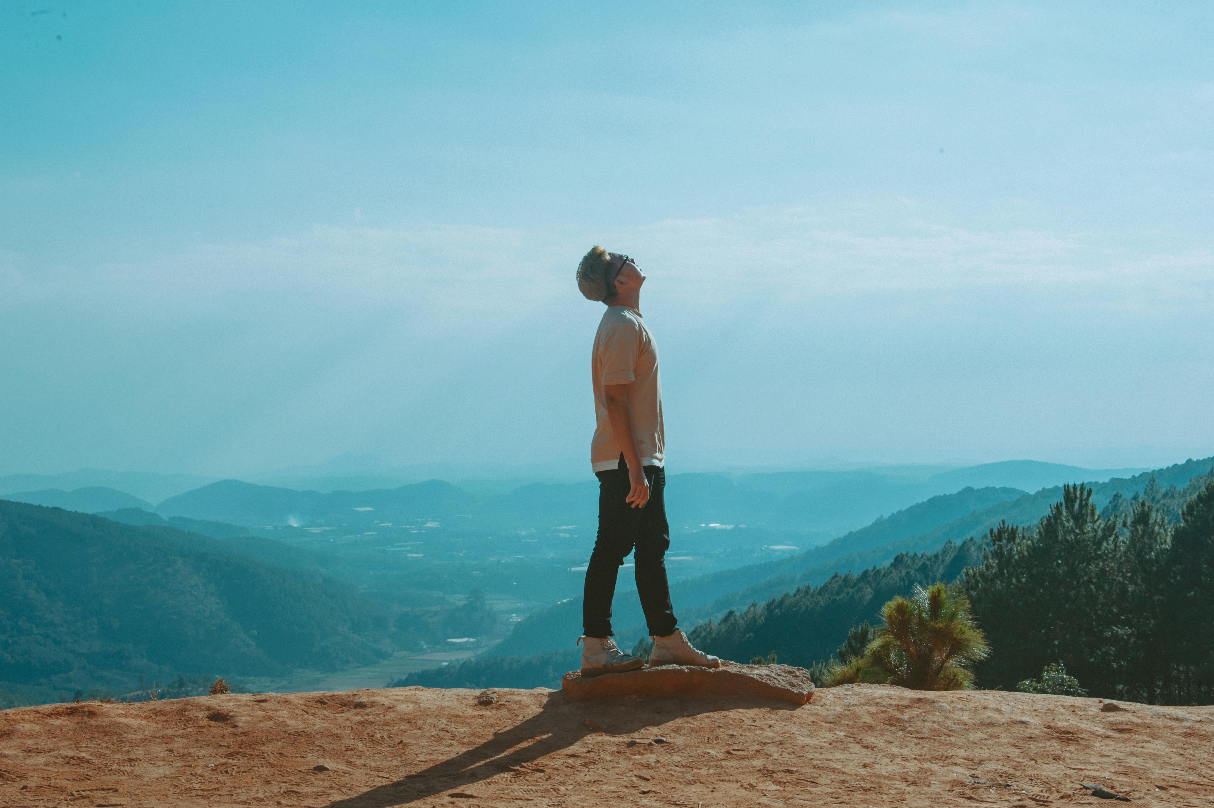 Free Man Wearing Beige Shirt and Black Pants Looking Up With Blue Sky and Mountains in the Background Stock Photo