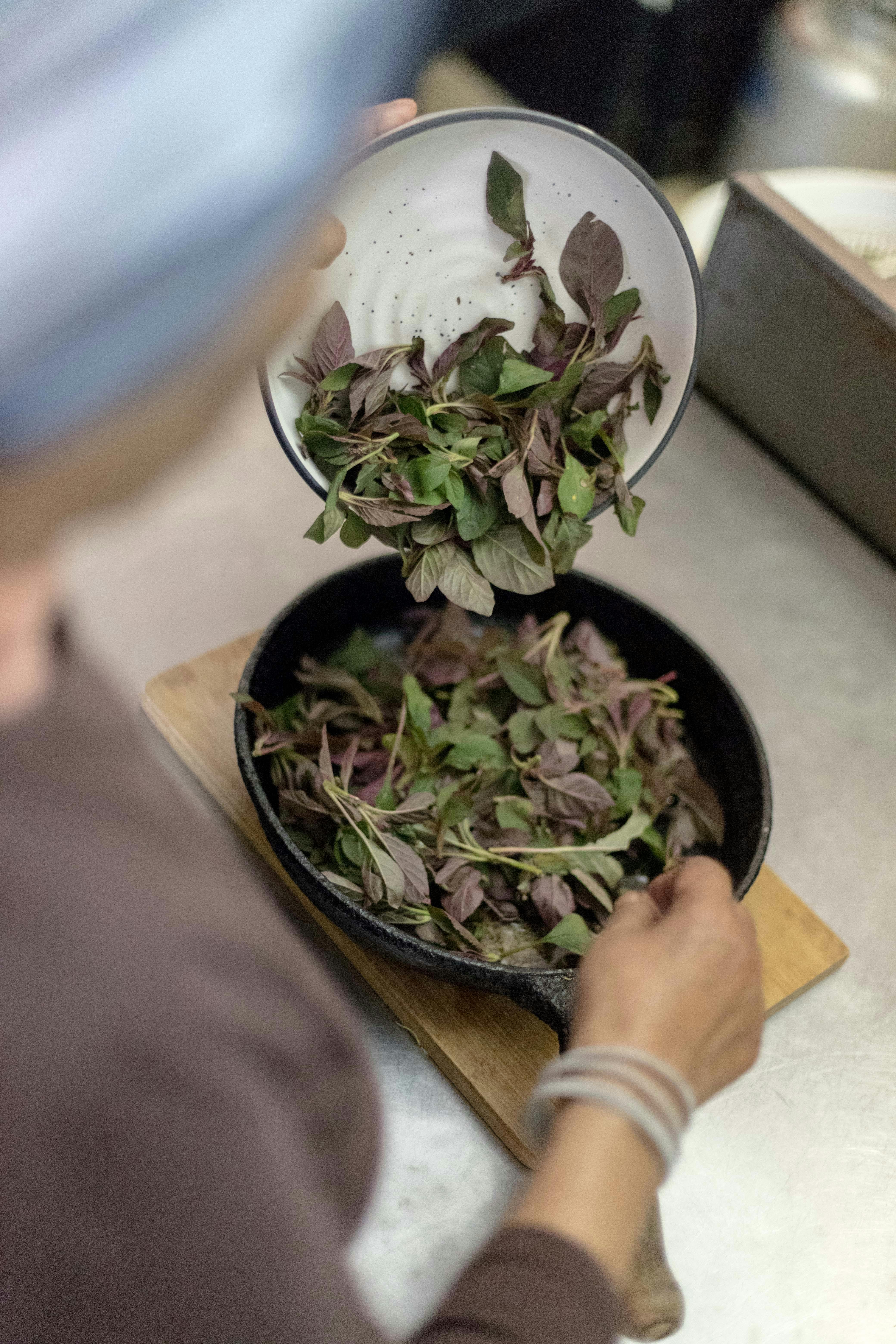A person adds fresh basil leaves to a cast iron skillet in a kitchen setting.