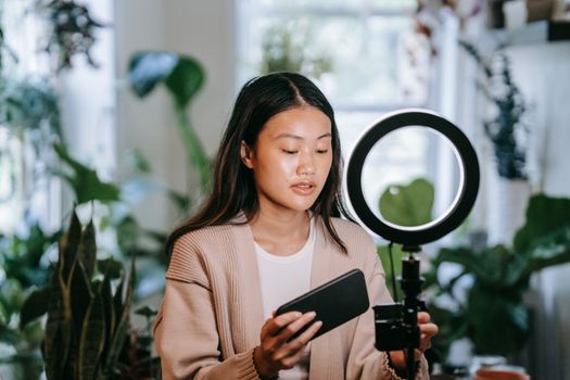 Asian woman adjusting smartphone and ring light indoors for video recording.
