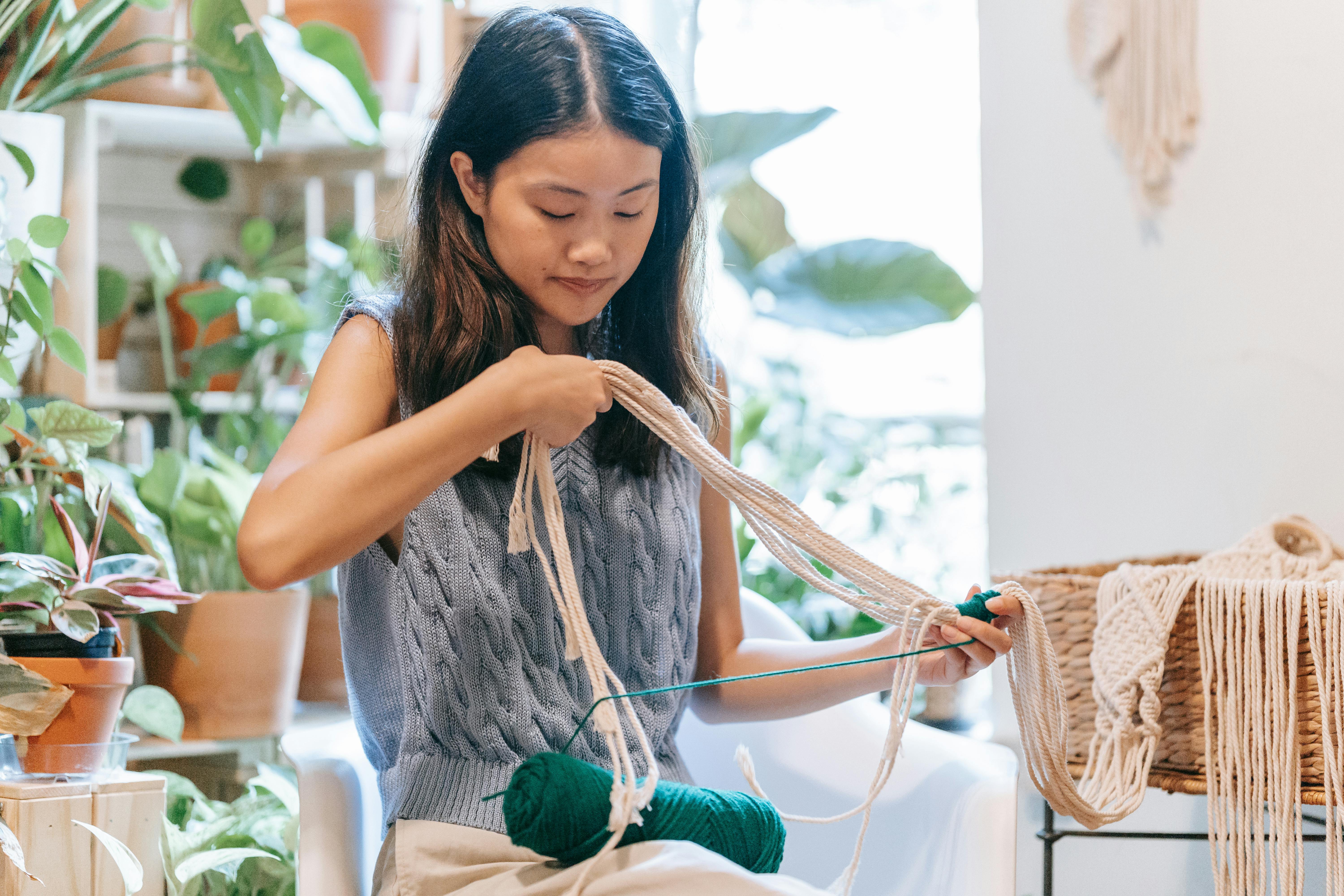 Asian woman skillfully crocheting with yarn in a comfortable indoor space surrounded by plants.