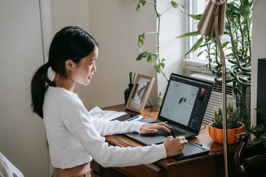 Teenage girl working on digital art with laptop and stylus in cozy home office setting.